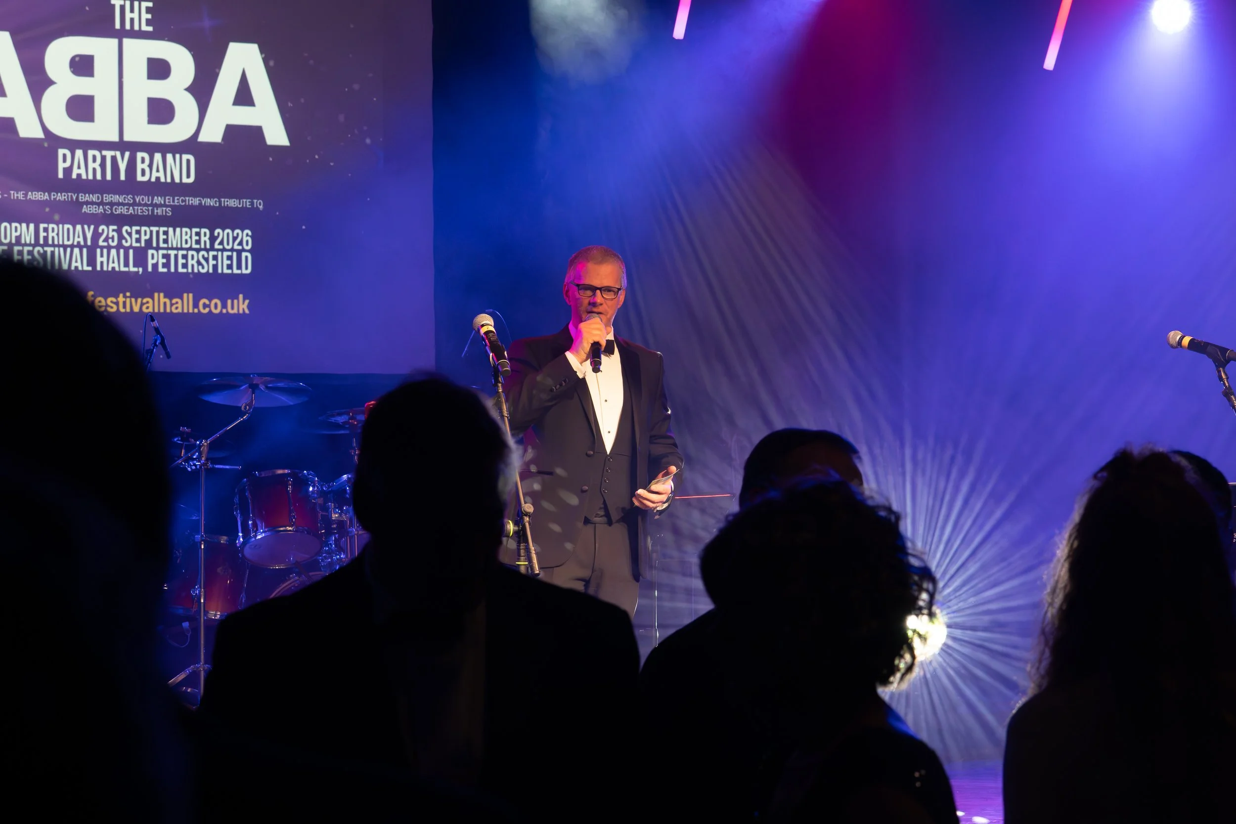 A man in a tuxedo with glasses speaking into a microphone on stage at a music event, with a drum set and a large screen behind him displaying an event poster for an ABBA tribute band.