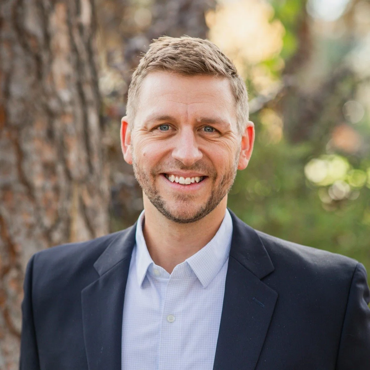 A smiling man with short blond hair and a beard, wearing a dark suit jacket and light blue shirt, standing outdoors near a tree with blurred green and brown background.