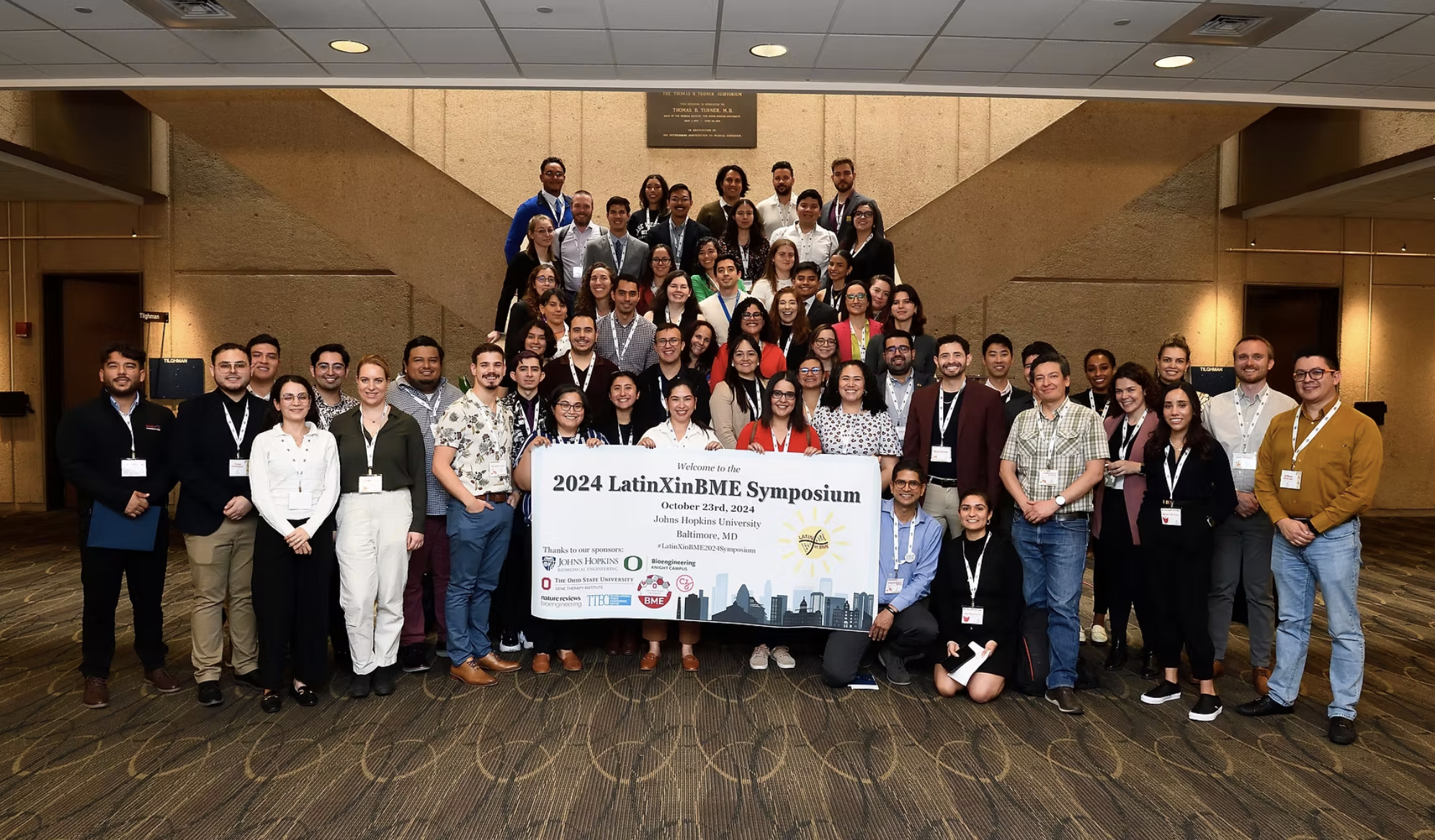 People at a symposium gathered around and holding a white banner with the words "2024 LatinXinBME group picture Symposium" on it