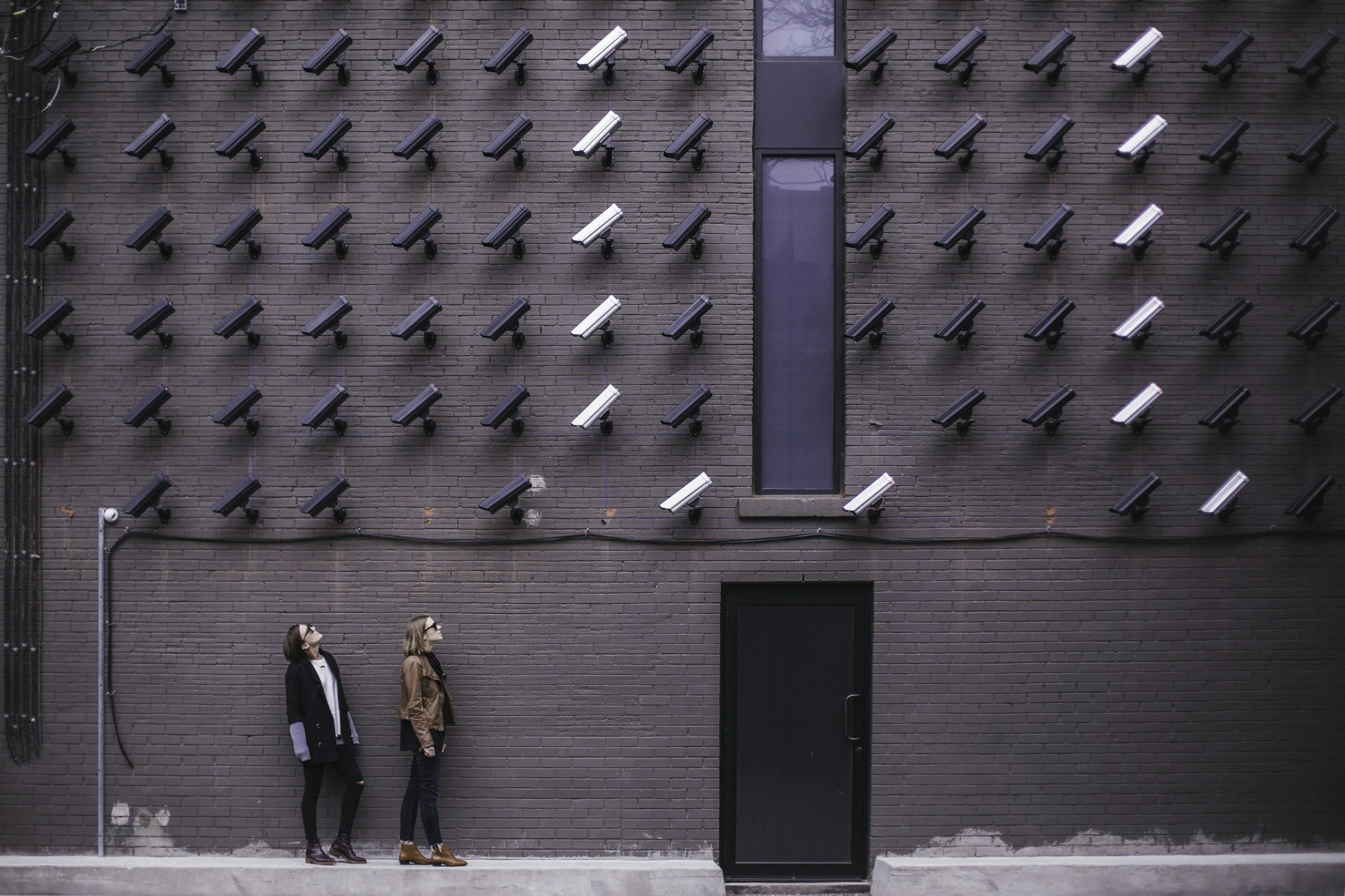A photo of two women standing in front of a building covered with security cameras pointing at them.