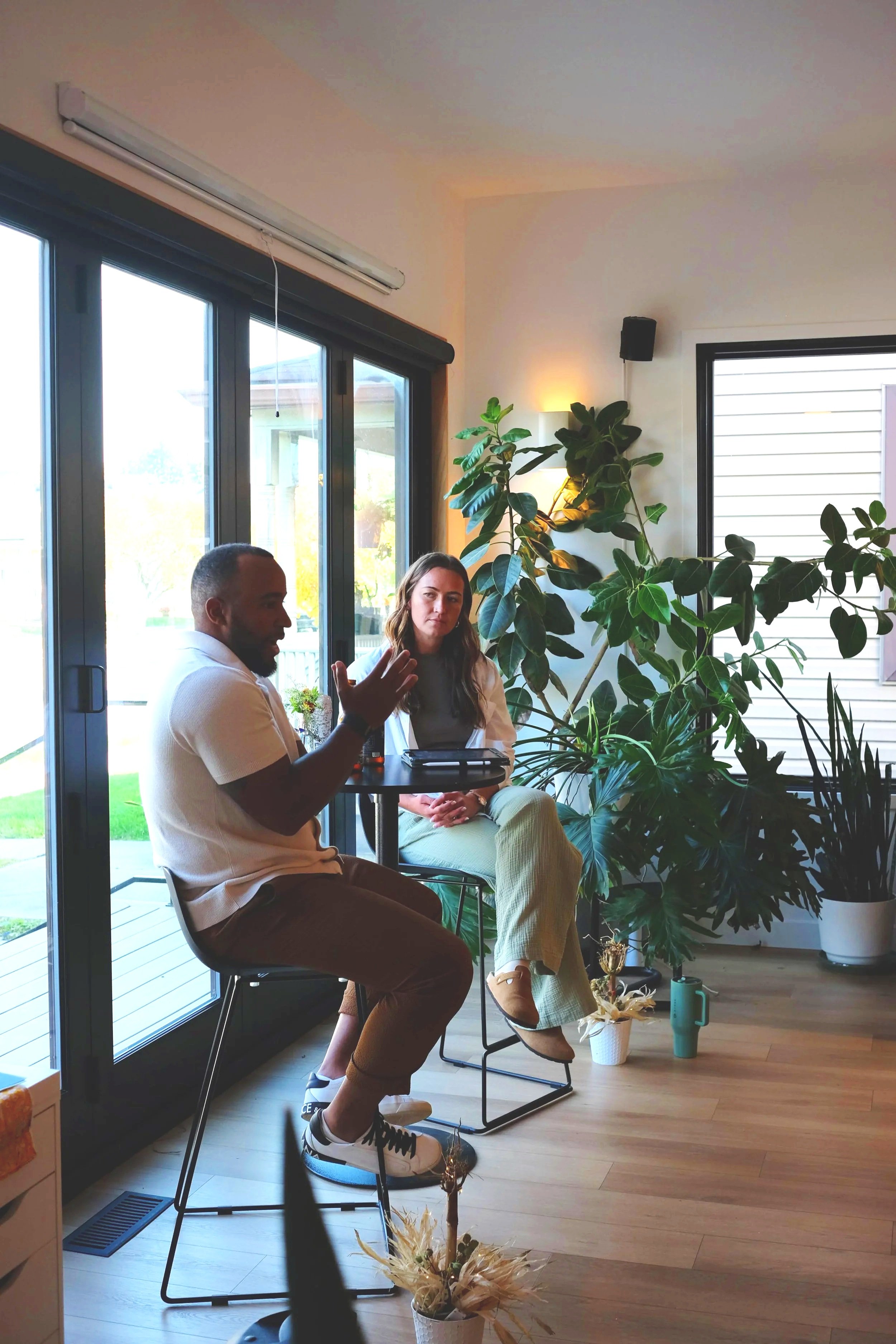 Two people sitting at a high table near large windows in a room filled with indoor plants. The man on the left is talking and gesturing, wearing a white polo shirt and brown pants. The woman on the right is listening, wearing a light-colored blazer and checkered pants. There are various plants, a watering can, a cup, and decorative items on the floor and window sills.