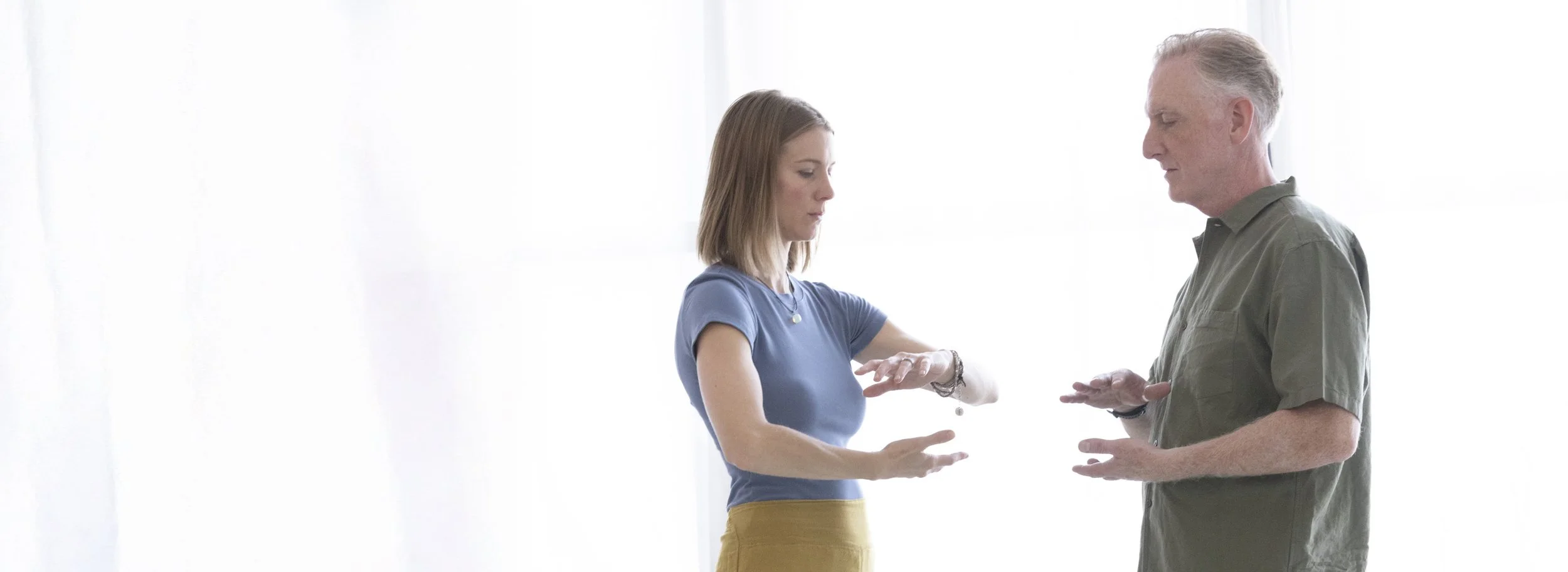 A woman and a man standing indoors, with the woman looking at her watch or bracelet and the man looking at her, both engaged in a conversation.