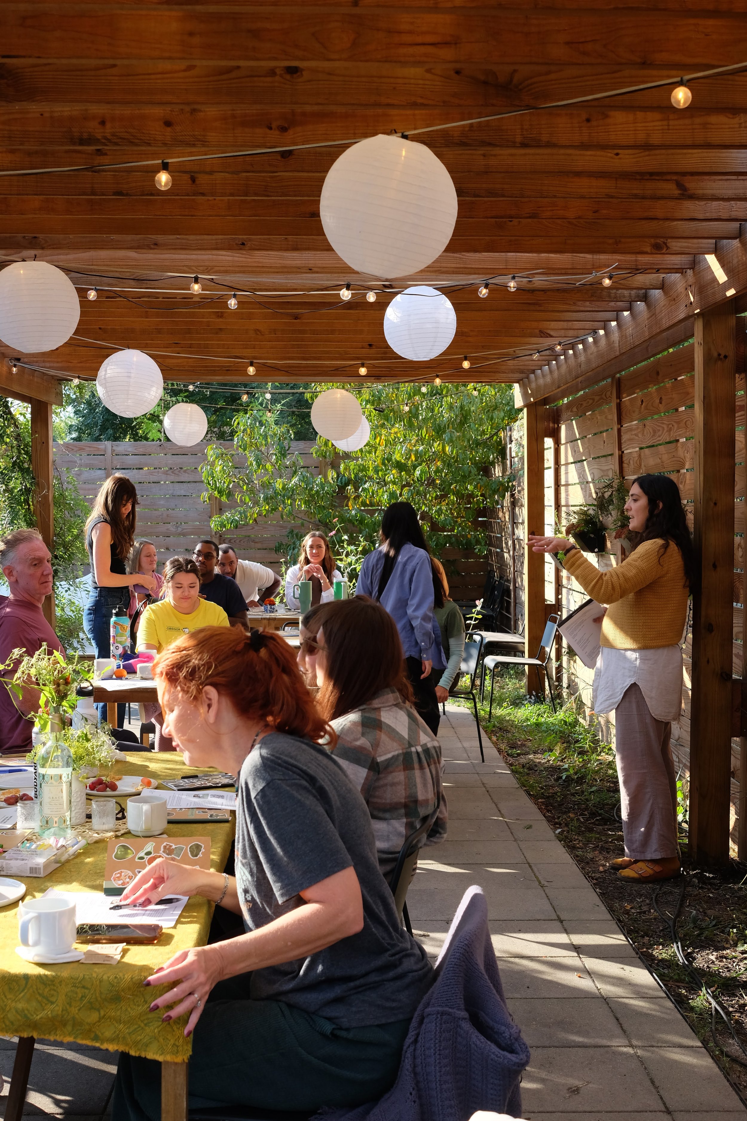 People gathered at an outdoor patio, some seated at a long table with snacks and drinks, and others standing and engaging in conversation, under hanging paper lanterns and string lights, with a wooden pergola and greenery in the background.