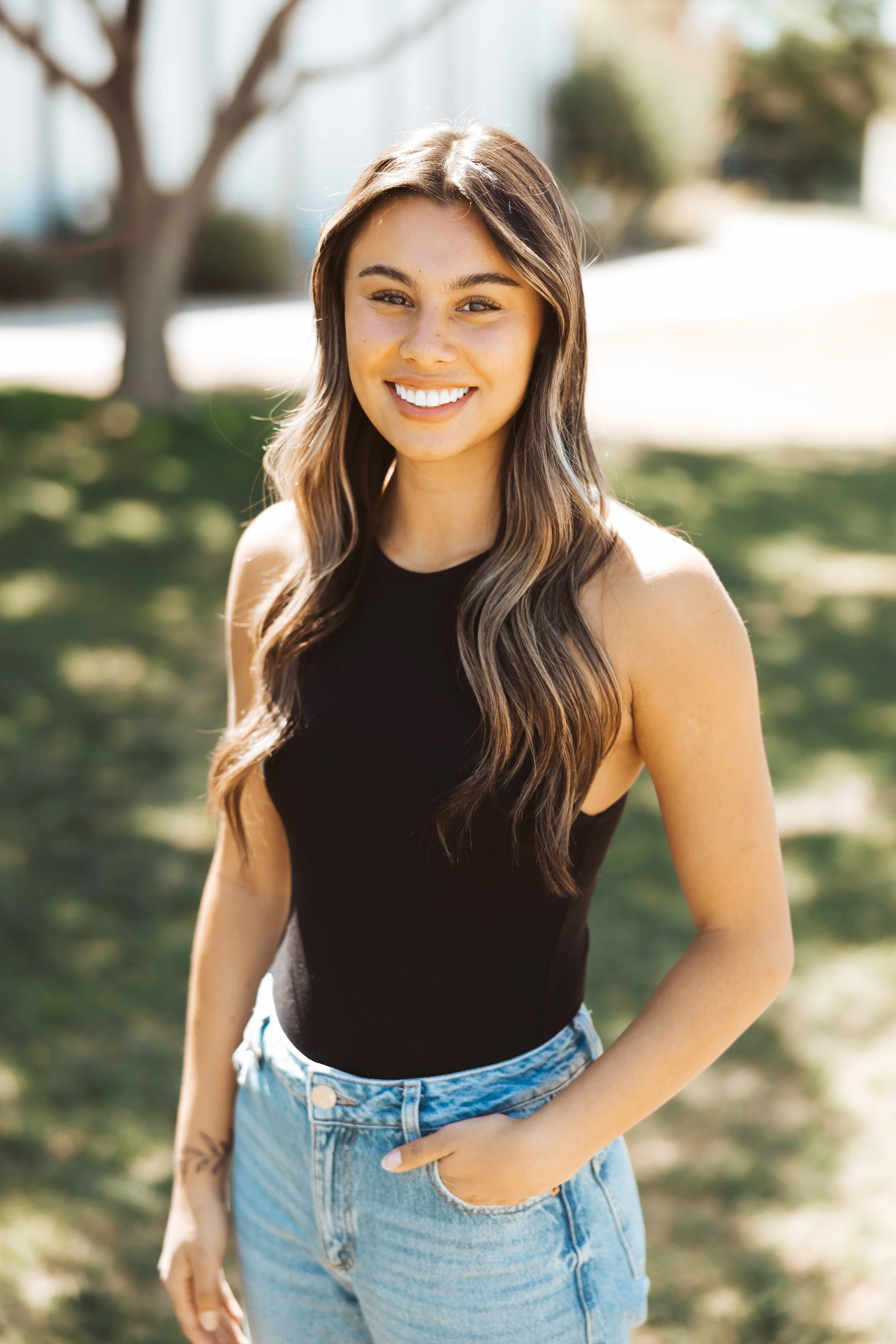 Young woman with long wavy brown hair smiling outdoors in sunlight, wearing a black sleeveless top and blue jeans.