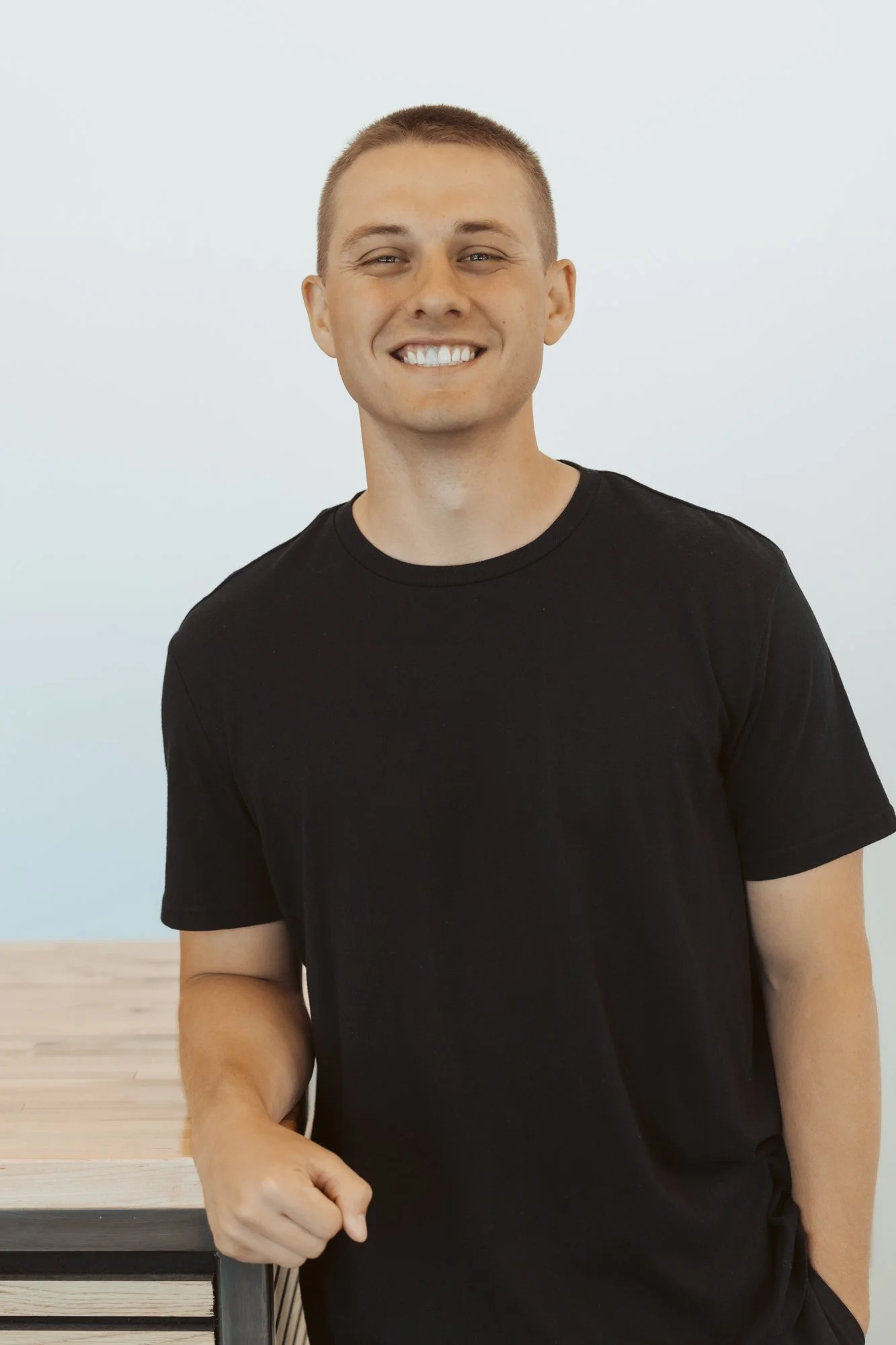 A young man with short blond hair, smiling, wearing a black t-shirt outdoors with trees and a building in the background.