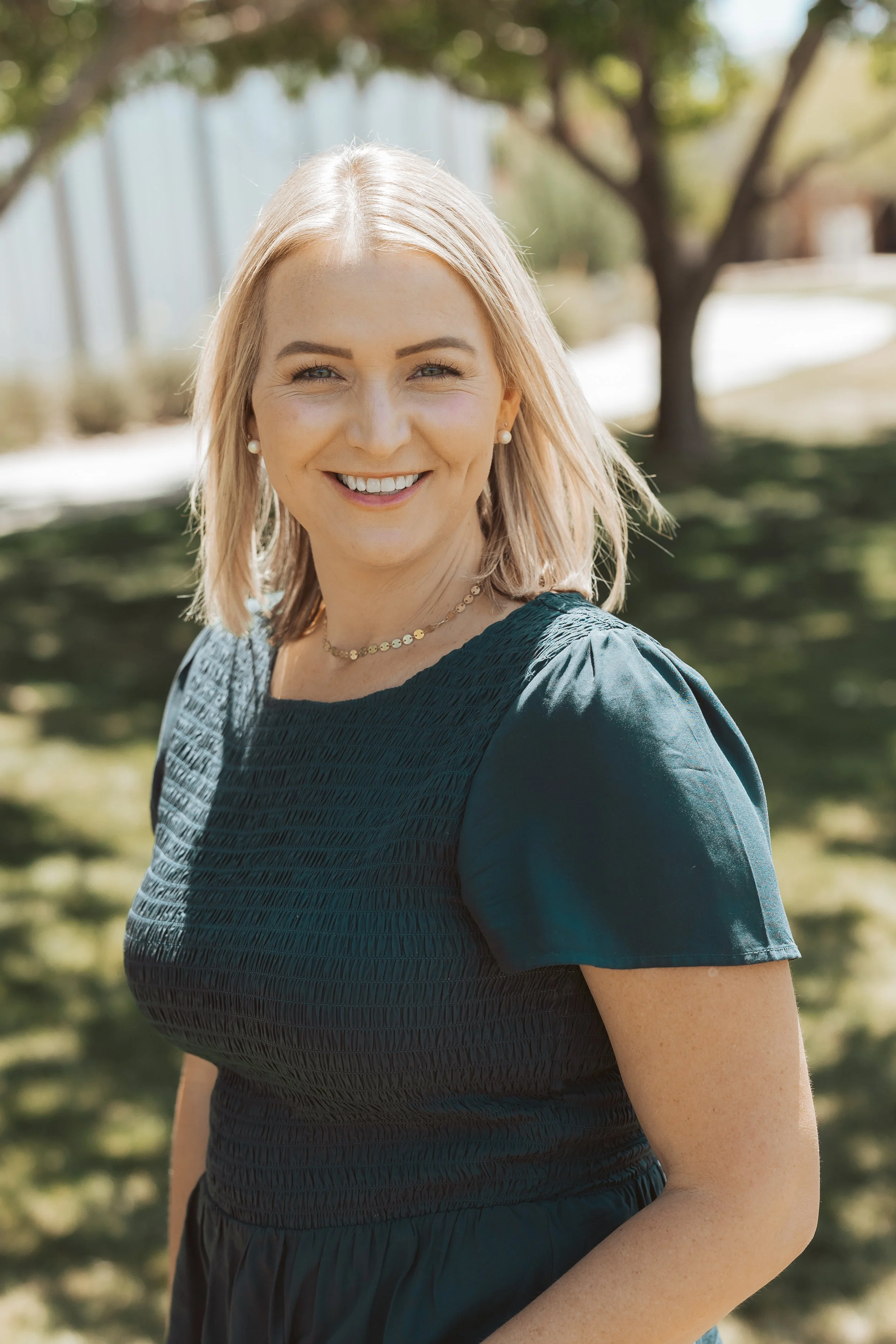 A smiling woman with blonde hair standing outdoors in sunlight, wearing pearl earrings, a necklace, and a dark dress with textured fabric and puffed sleeves.