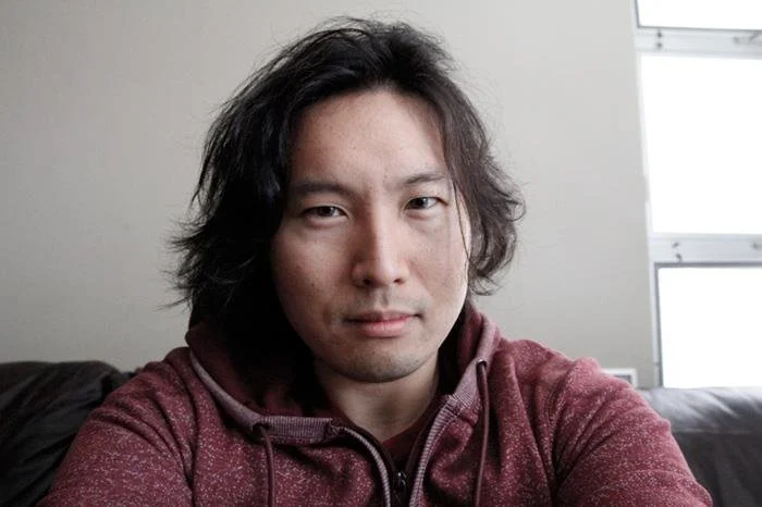 A man with medium-length dark hair and light skin taking a selfie indoors, wearing a maroon hoodie.