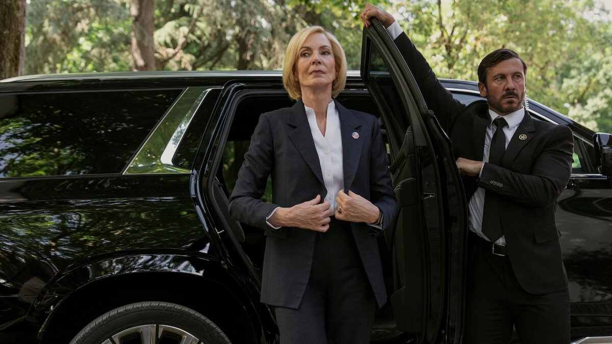 A woman stepping out of an SUV pulling her suit jacket tight to her chest as a man in a suit holds the door open