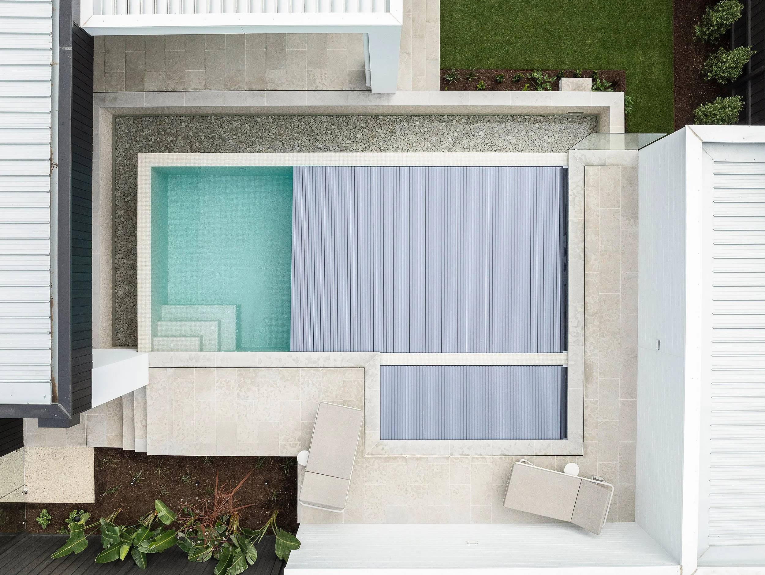 Aerial view of a modern backyard pool area with a covered section, surrounded by patio, small garden, and neighboring structures.