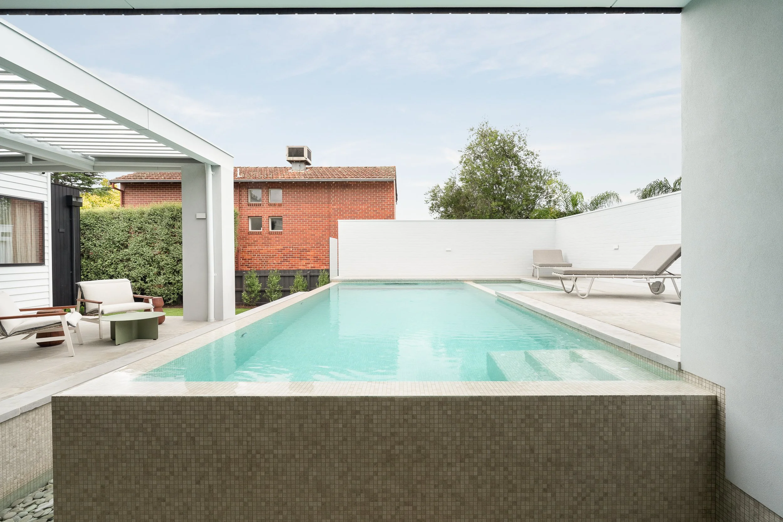 A private backyard swimming pool with lounge chairs and outdoor furniture, enclosed by a white brick wall and surrounded by greenery and neighboring buildings.