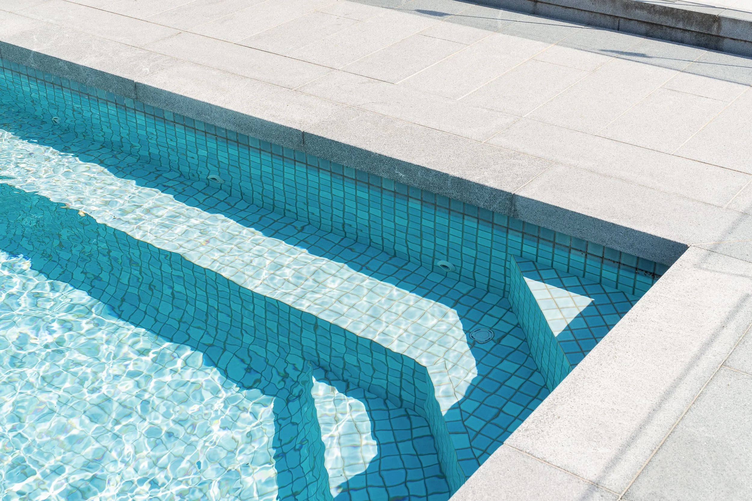 Close-up of a swimming pool with clear blue water and tiled steps, surrounded by a concrete deck.