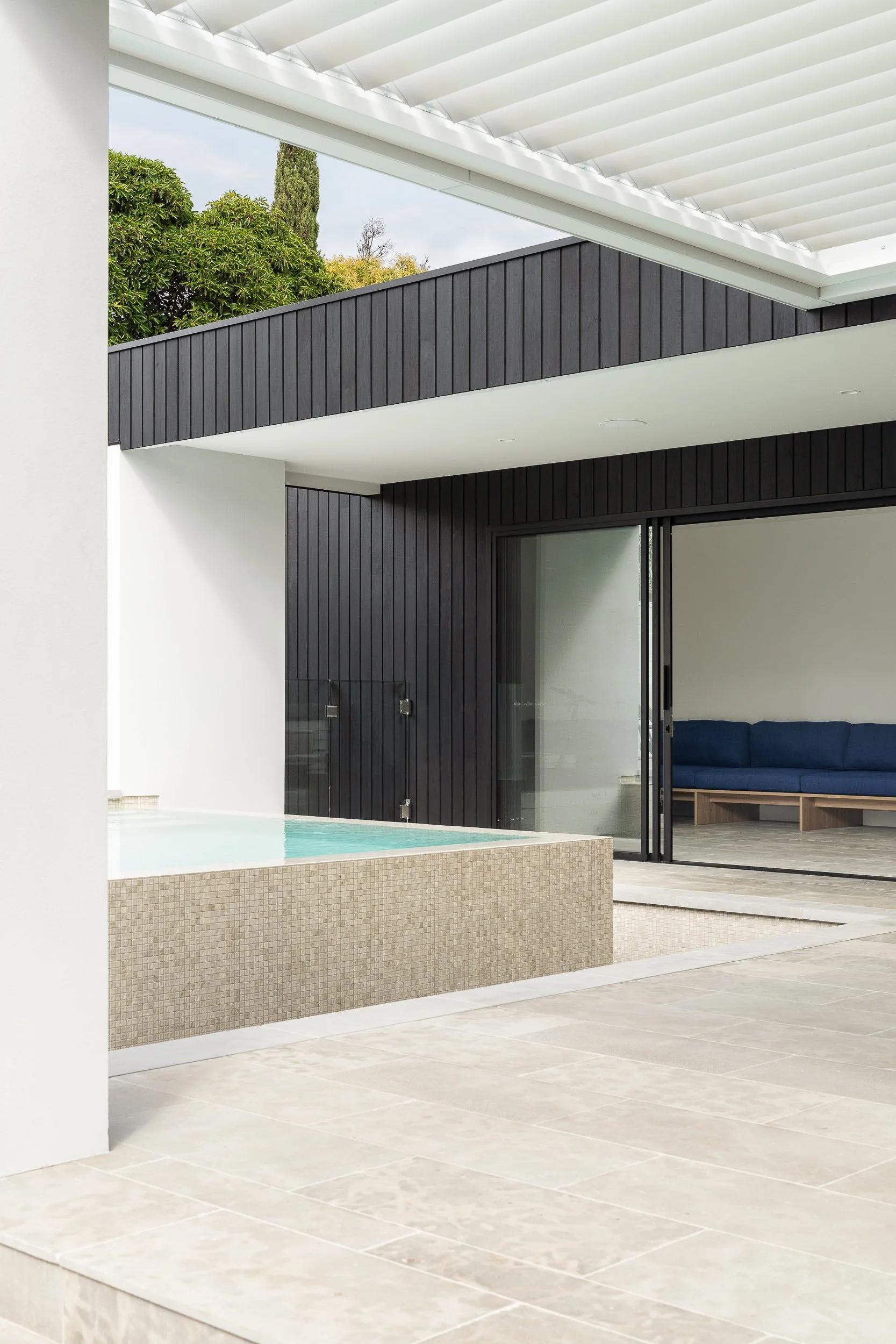 Modern outdoor patio area with a small pool, light-colored tiled floor, black wooden wall, and sliding glass door leading to an indoor room with a blue sofa.