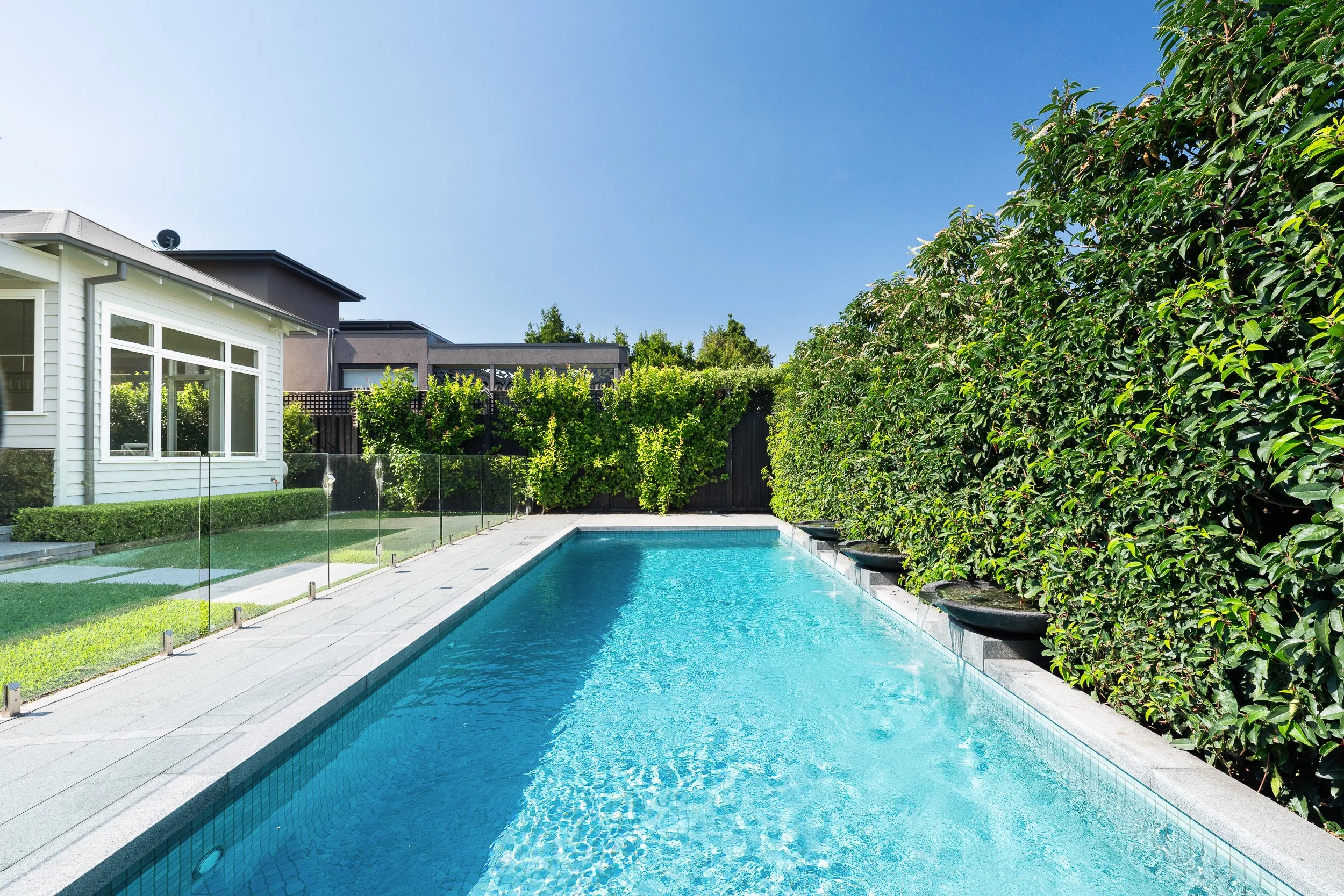 A backyard swimming pool with clear blue water, surrounded by green bushes and trees, with a white house and sky in the background.