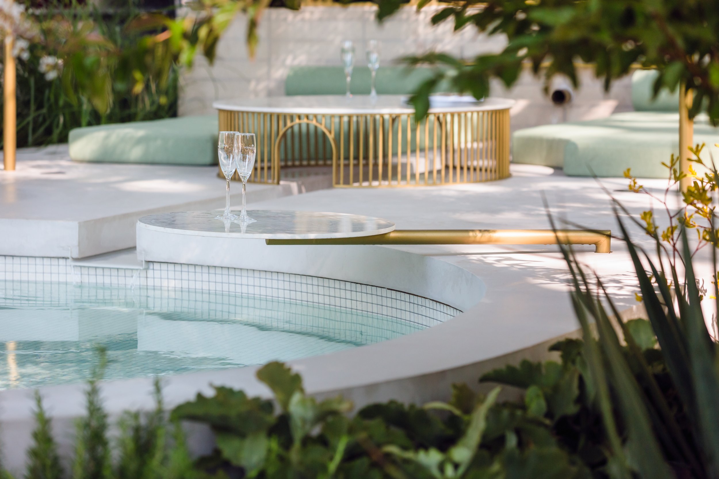A backyard hot tub with two champagne glasses on a white marble side table, surrounded by lush green plants and outdoor seating with green cushions. There is another set of champagne glasses on a golden round table in the background.
