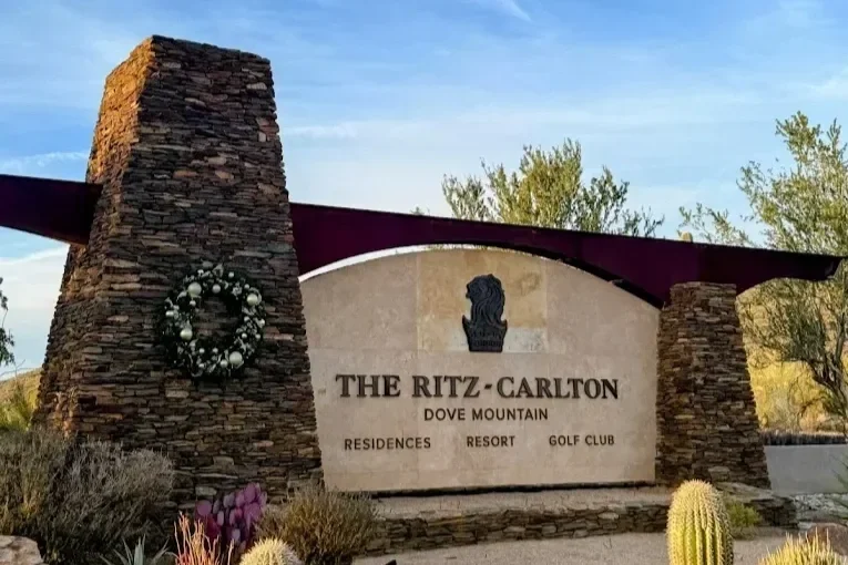 Sign for The Ritz-Carlton Dove Mountain resort, featuring a stone and metal structure, floral wreath, and desert plants in the foreground.