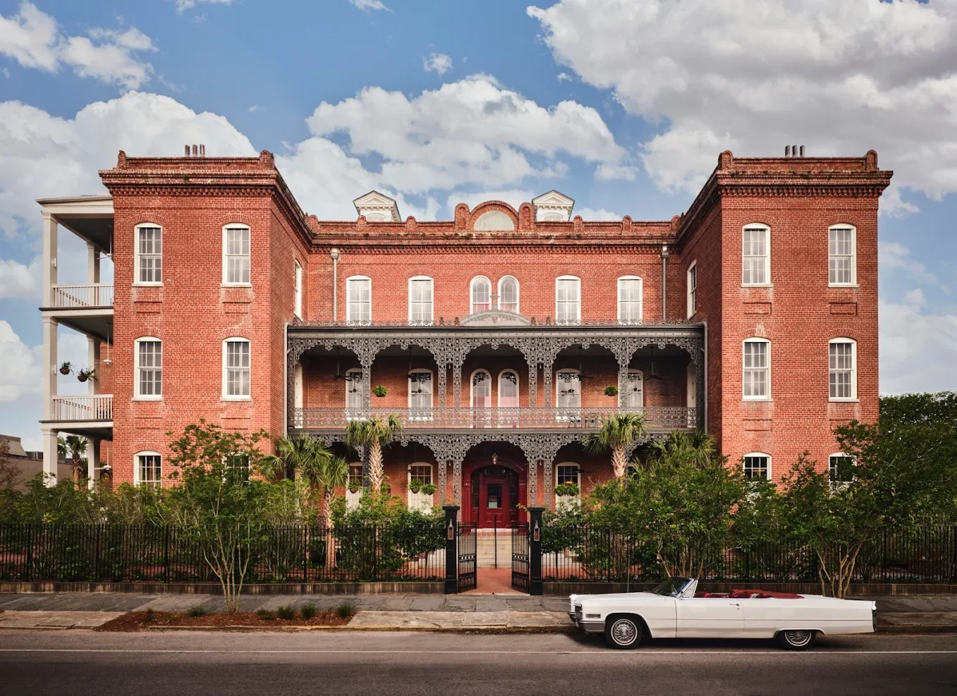 A large historic brick building with decorative ironwork balconies, surrounded by greenery and a black iron fence, with a white vintage car parked on the street in front.