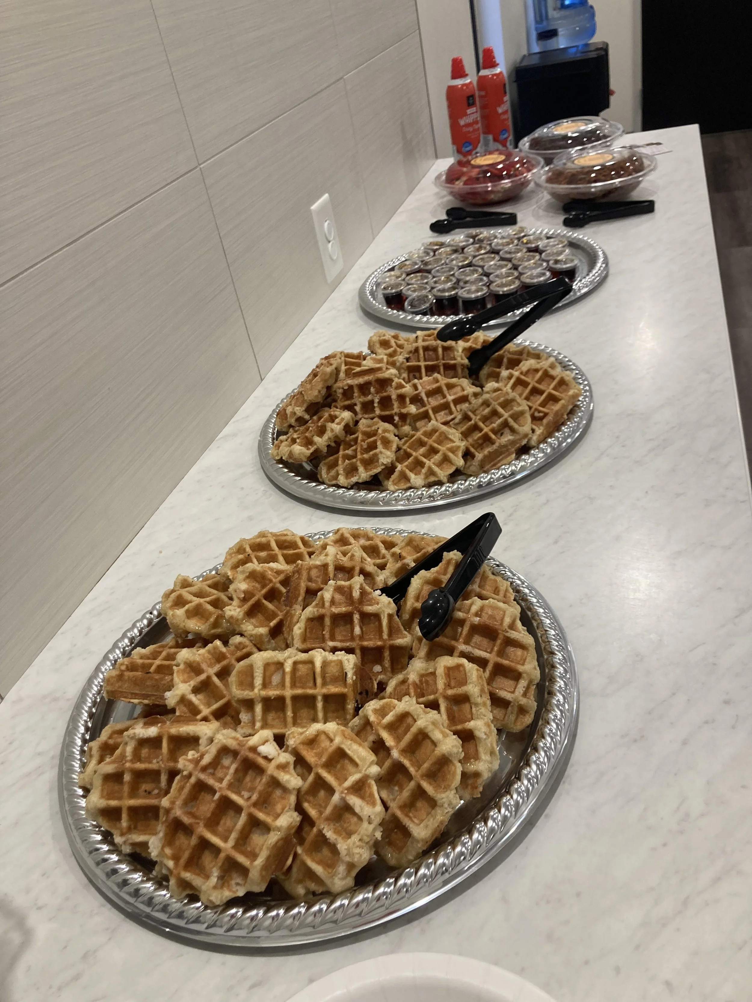 Table with trays of waffles, small containers of syrup, and other breakfast items in a cafeteria or kitchen setting.