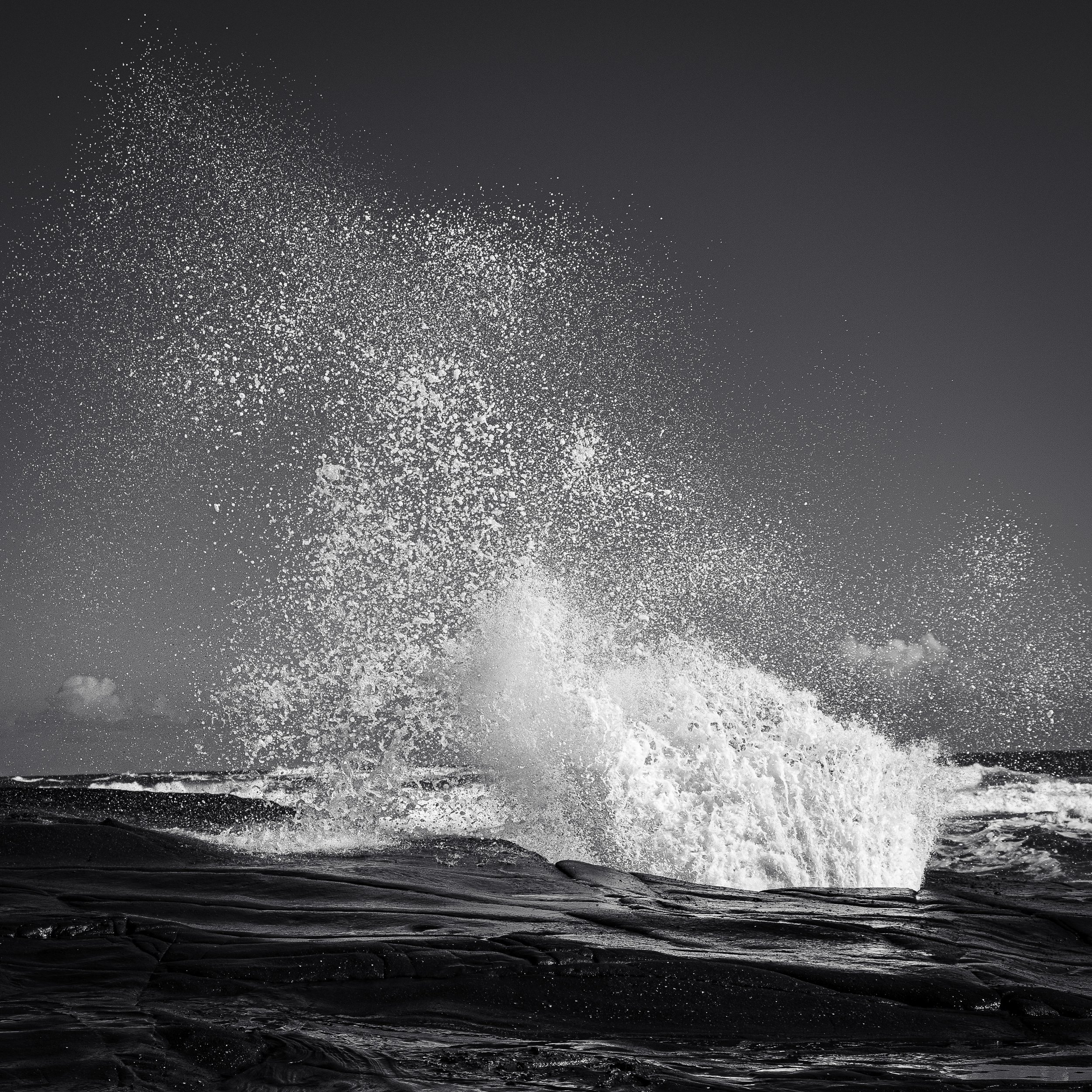 Black and white photo of a wave crashing against rocks, creating splashes and droplets against a clear sky.