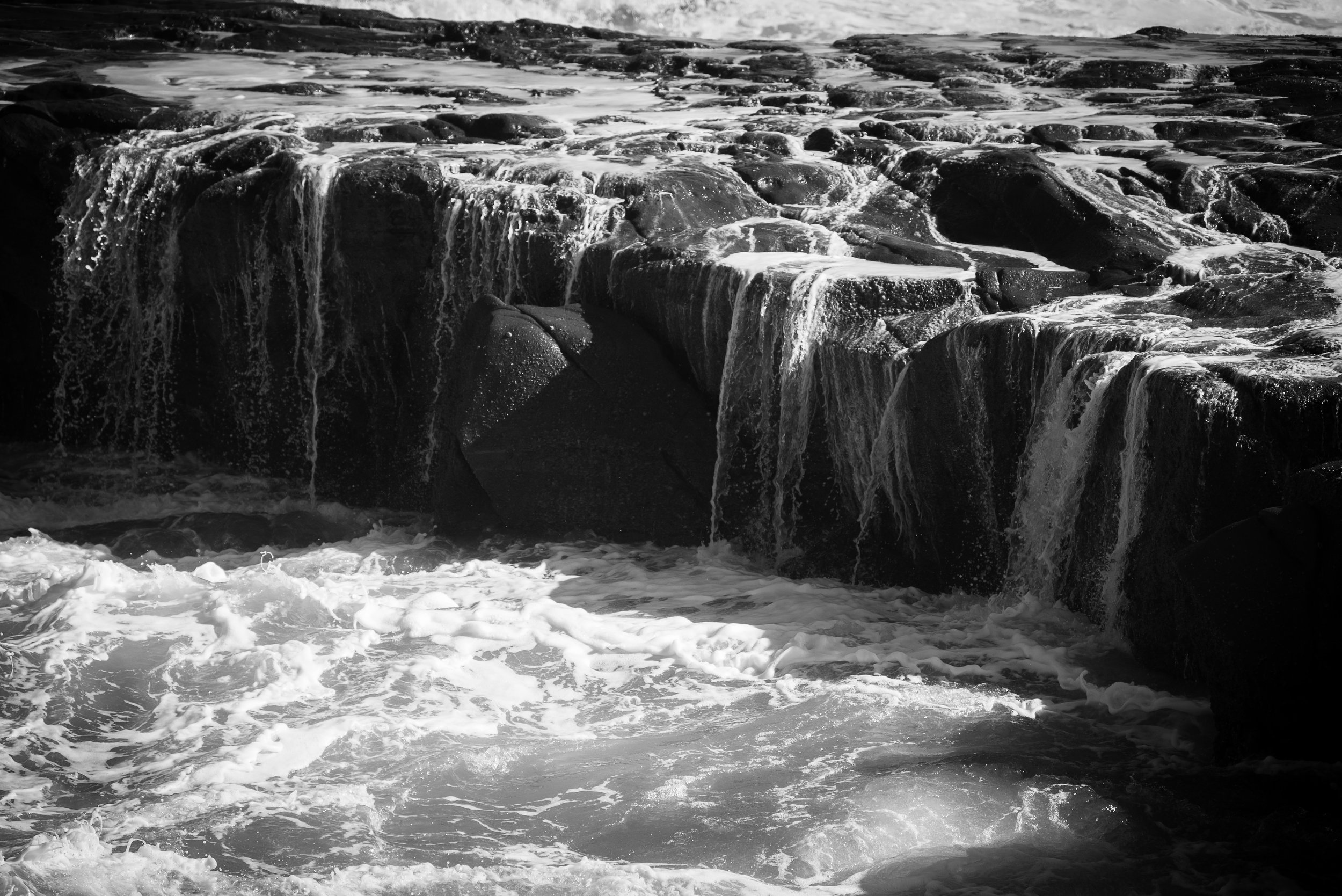 Black and white image of waves cascading over a rocky shoreline with water forming small waterfalls.