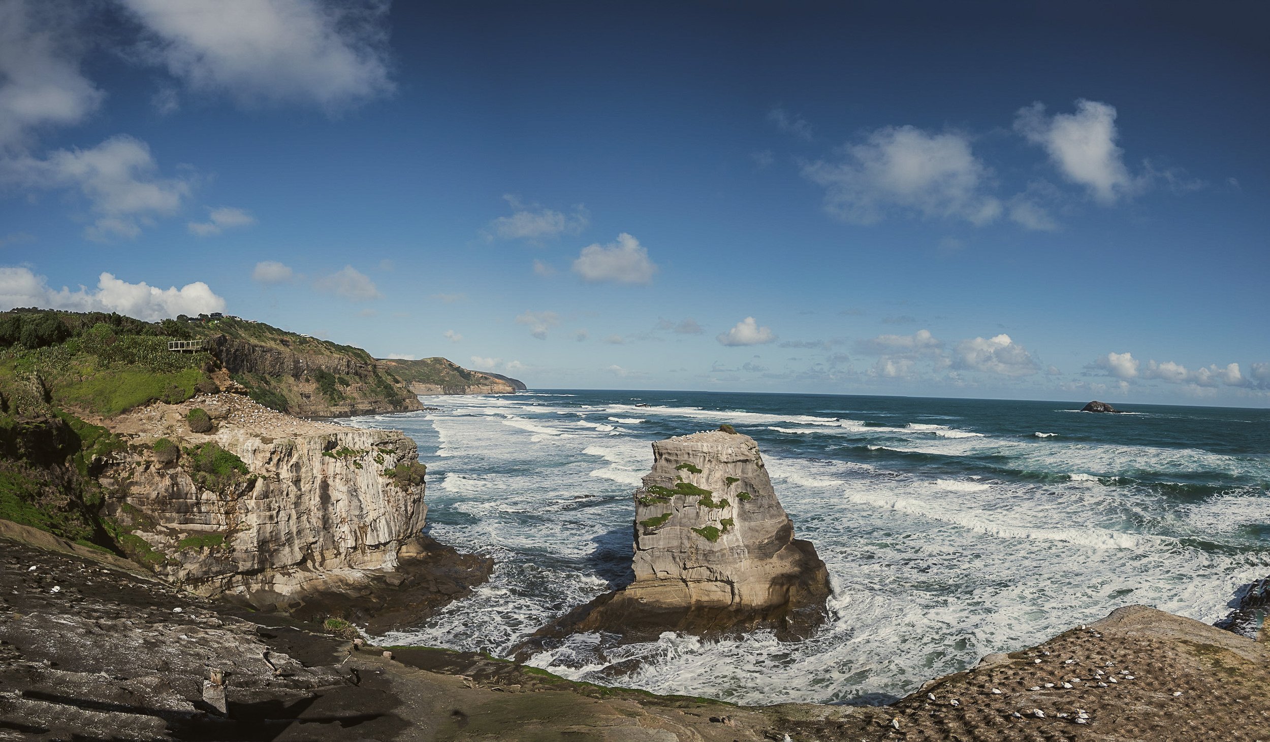 Scenic coastal view with cliffs, rock formations, and ocean waves under a blue sky with clouds.