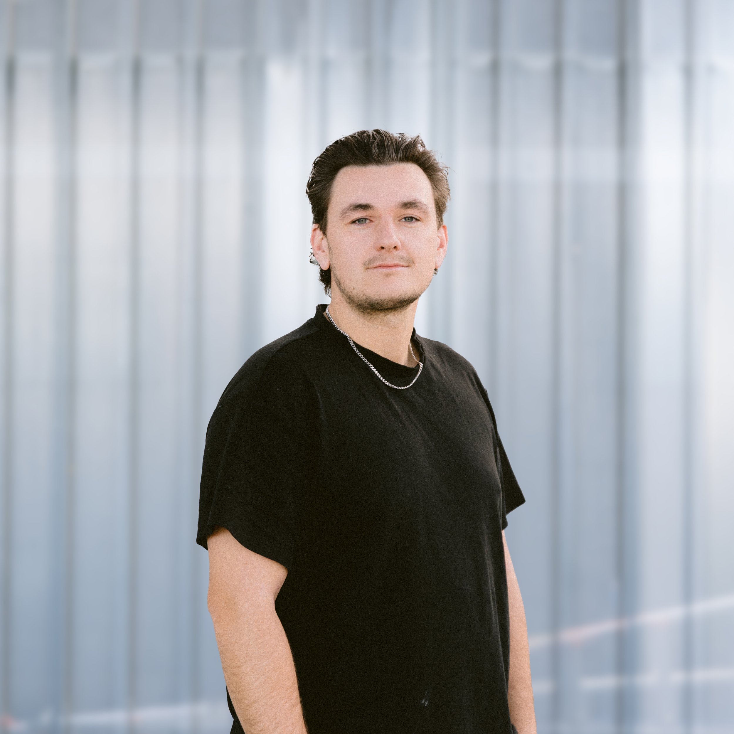 Portrait of a young man smiling, wearing a blue hoodie under a plaid jacket, with a light-colored backdrop