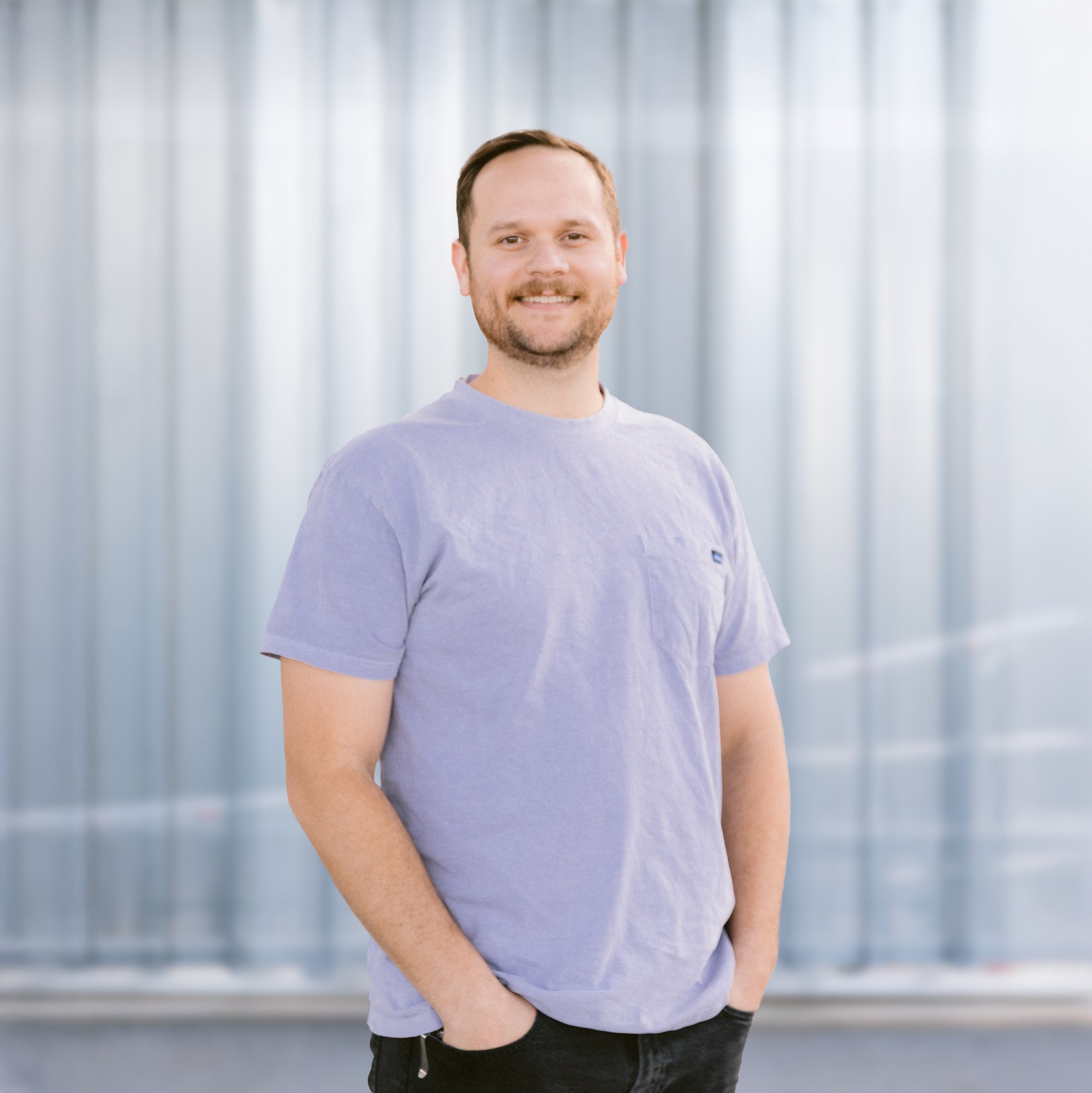 A smiling man with short brown hair, a beard, and blue eyes, wearing a blue button-up shirt, standing in front of a metal wall.