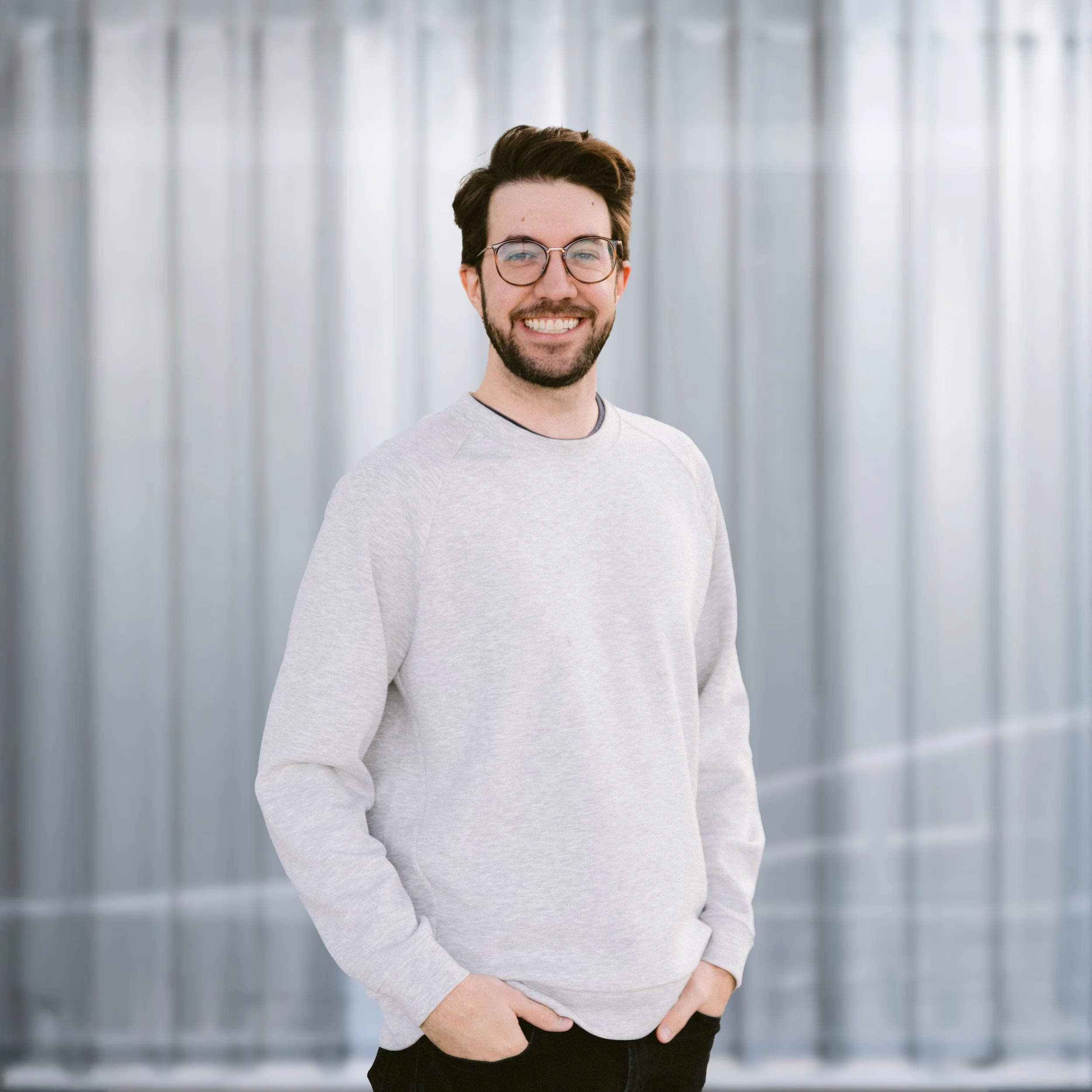 A smiling young man with dark hair, glasses, and a beard, wearing a black shirt, standing in front of a light-colored, vertically striped background.