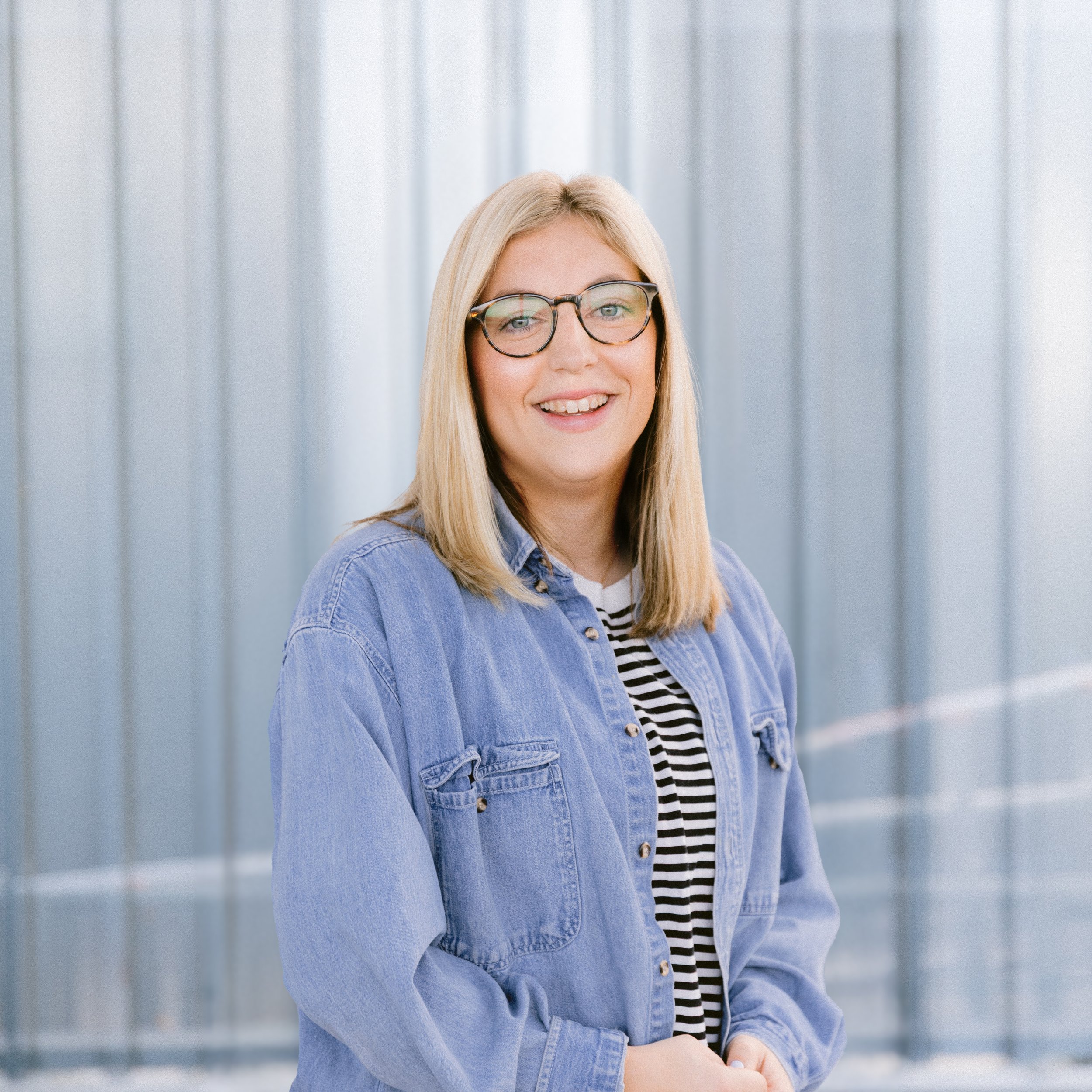 A young woman with long blond hair smiling at the camera, wearing a black shirt and a delicate necklace, in front of a glass wall with vertical lines.