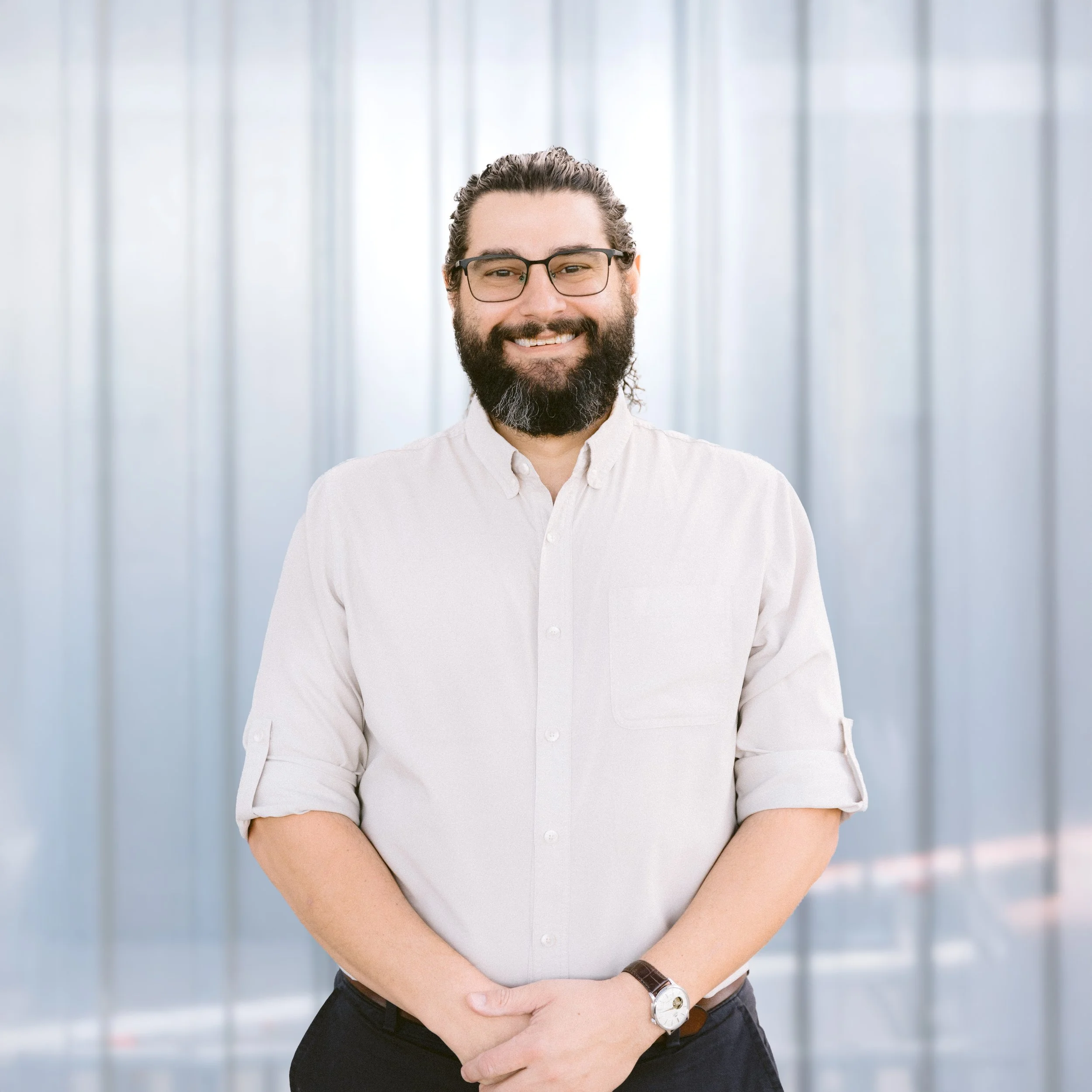 Smiling man with long hair tied back and a full beard, wearing a light blue button-up shirt, against a neutral background.