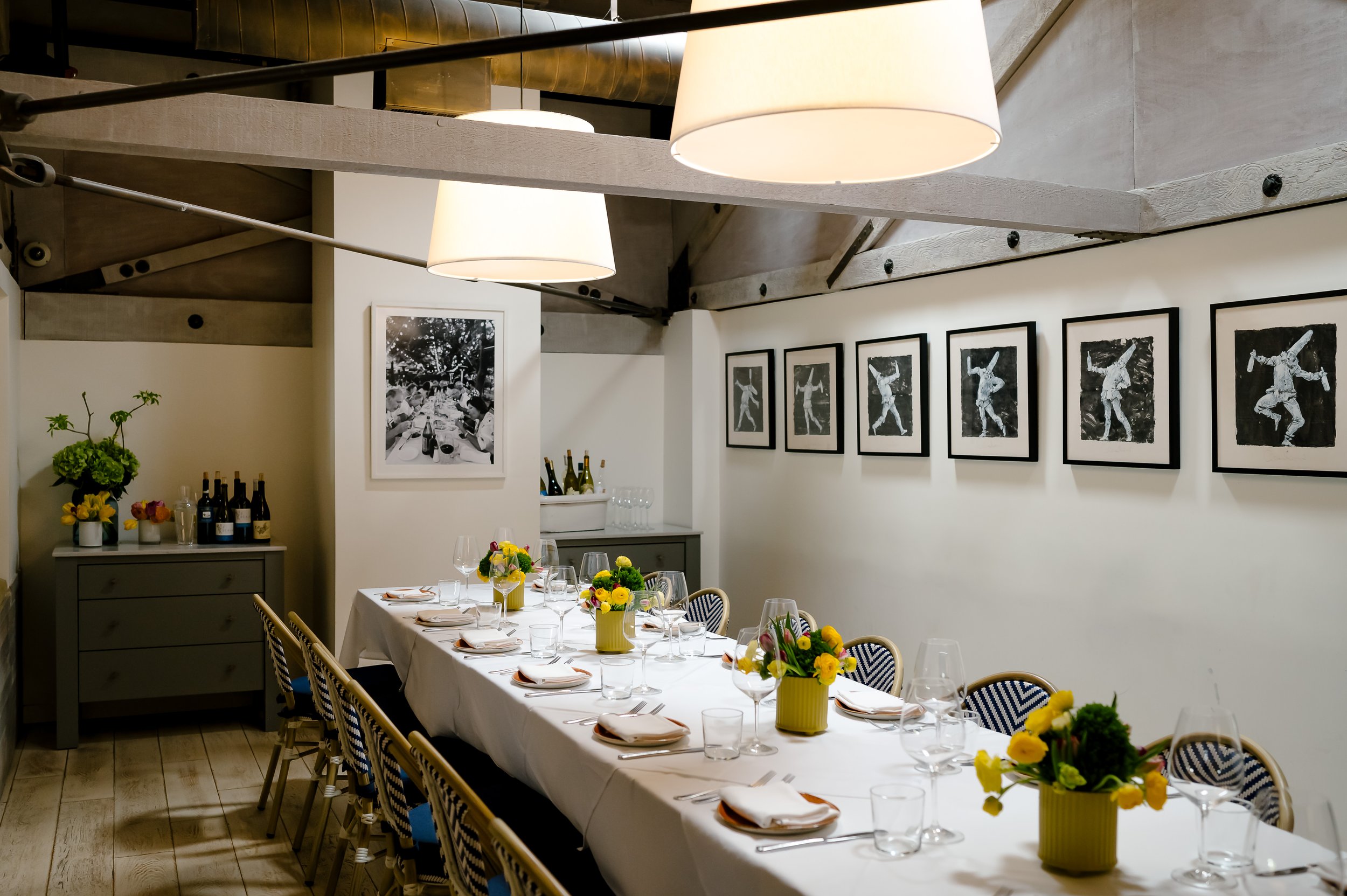 Private dining room at Scala Osteria in Napa Valley with a long table set for a group, white tablecloth, wine glasses, yellow floral arrangements, and framed artwork, ideal for intimate events and group dining.