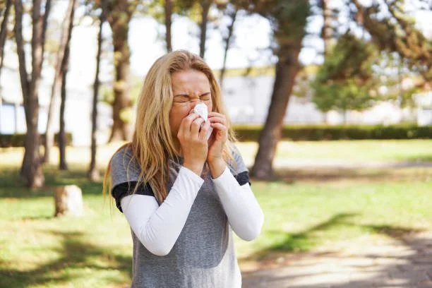 A picture of a blonde woman in a park sneezing and holding a tissue to her nose.