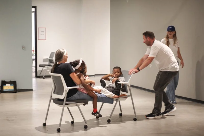 A man playfully interacts with young children sitting on chairs in a room with plain walls, while a woman and girl watch and smile, and a girl in a cap stands in the background.