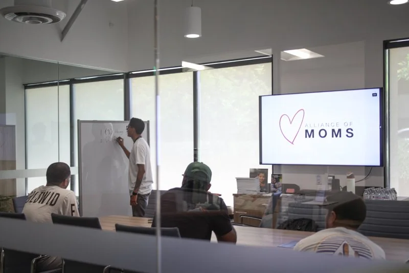 A classroom or meeting room with a man standing at a whiteboard and four people sitting at a table, with a large screen displaying 'Alliance of Moms' and a heart logo.