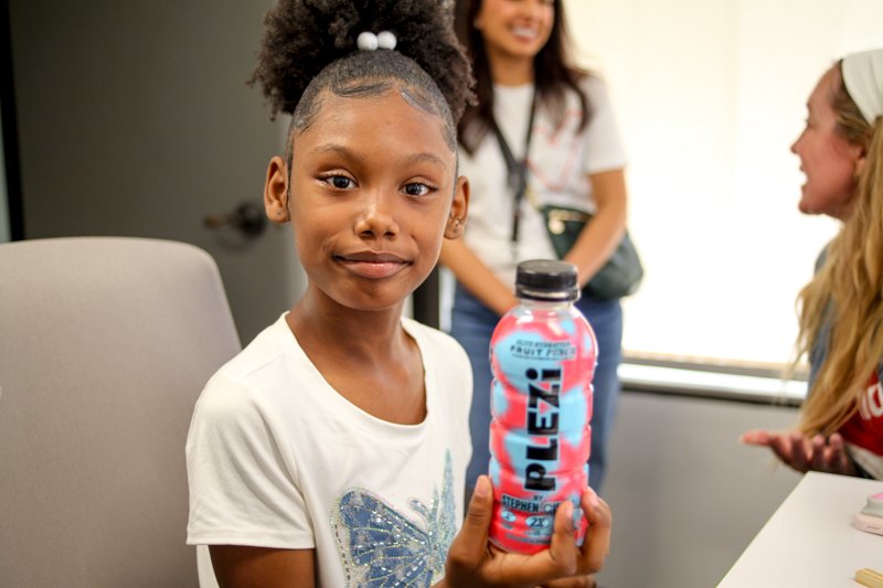 A young girl with curly hair in a ponytail holding a bottle of Lez Hydrate flavored water, with two women in the background smiling and chatting in a bright room.