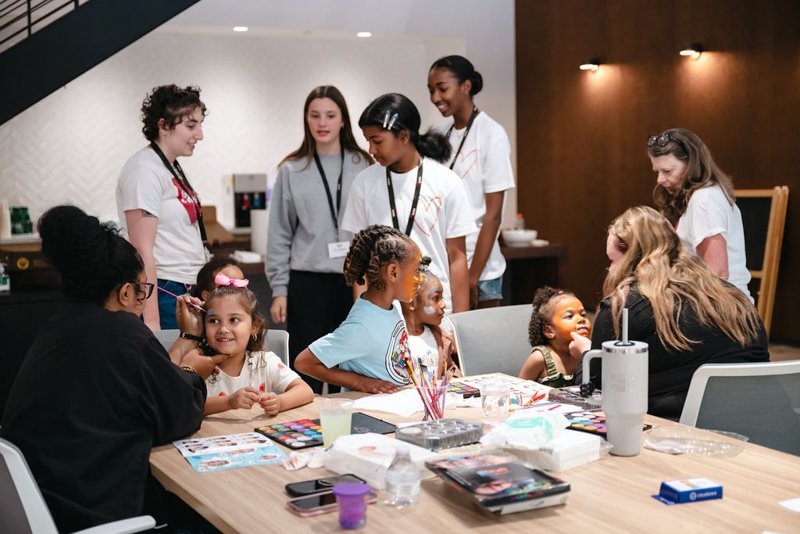 Children and adults in a room engaged in face painting activity at a table with art supplies, some standing and some sitting, in a casual indoor setting.
