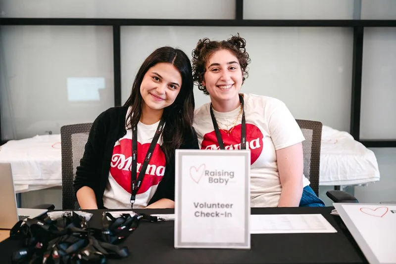 Two women sitting at a table with a sign that reads 'Raising Baby Volunteer Check-In,' wearing matching 'Diana' shirts with red hearts, smiling at the camera.