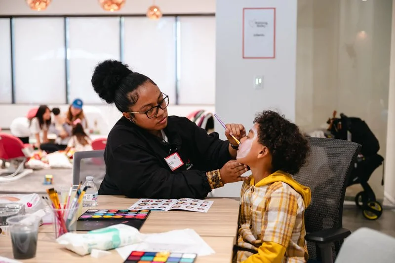 A woman with glasses and a black hoodie is applying face paint to a young boy with curly hair, wearing a yellow plaid shirt, in a vibrant indoor setting. There are art supplies and a water bottle on the table, with other children and adults in the ba