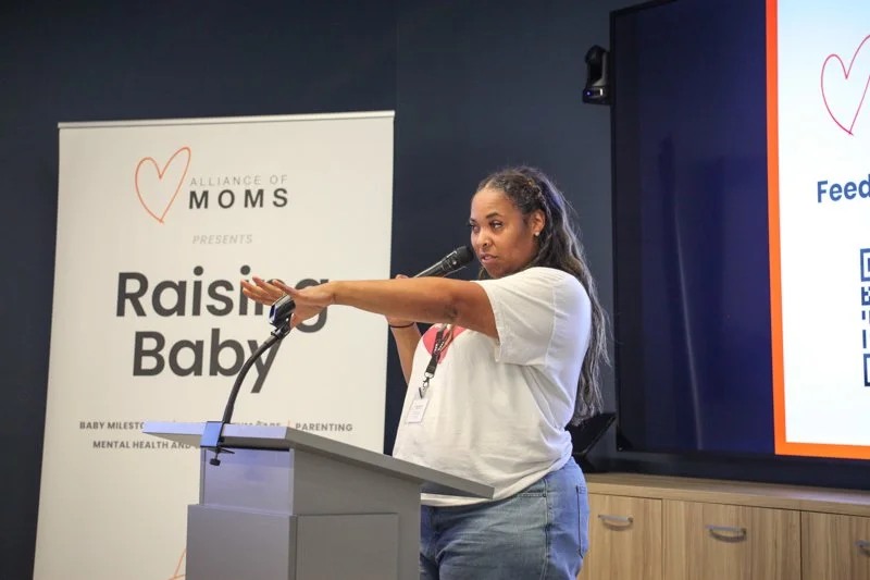 A woman speaking at a podium during a presentation at an event called 'Raising Baby,' organized by the Alliance of Moms, with a banner displaying the event name and a screen with additional information.