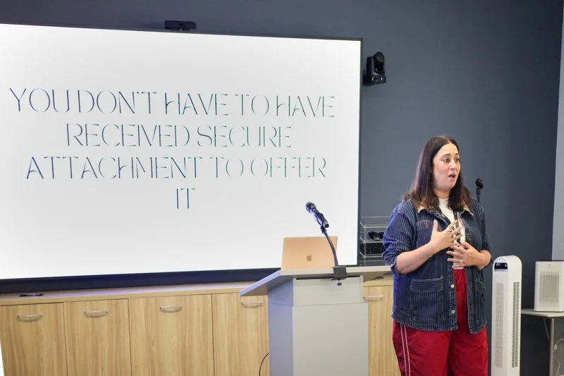 A woman in a denim jacket and red pants giving a presentation in a room with a large screen behind her. The screen displays a quote about attachment and security.
