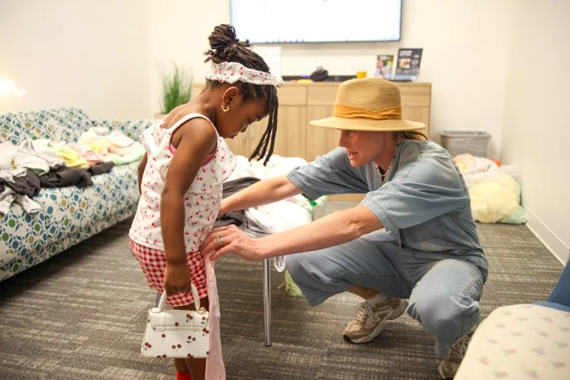 A woman in a wide-brimmed hat and casual clothes crouches down to interact with a young girl holding a small purse in a room with laundry on a couch and a TV in the background.