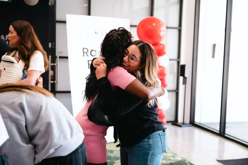 Two women hugging and smiling in a bright, modern lobby with red balloons and a sign in the background.