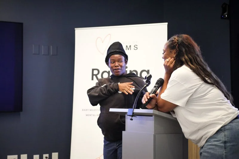 A young girl on stage speaking into a microphone, with a woman leaning over a podium next to her, against a background with a banner displaying the words 'Raising' and a heart symbol.
