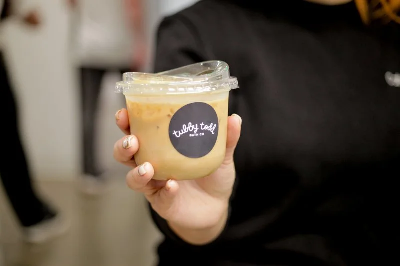 A person holding a clear plastic container of a creamy, beige-colored substance with a black circular label that reads "tubby todd BATH CO".