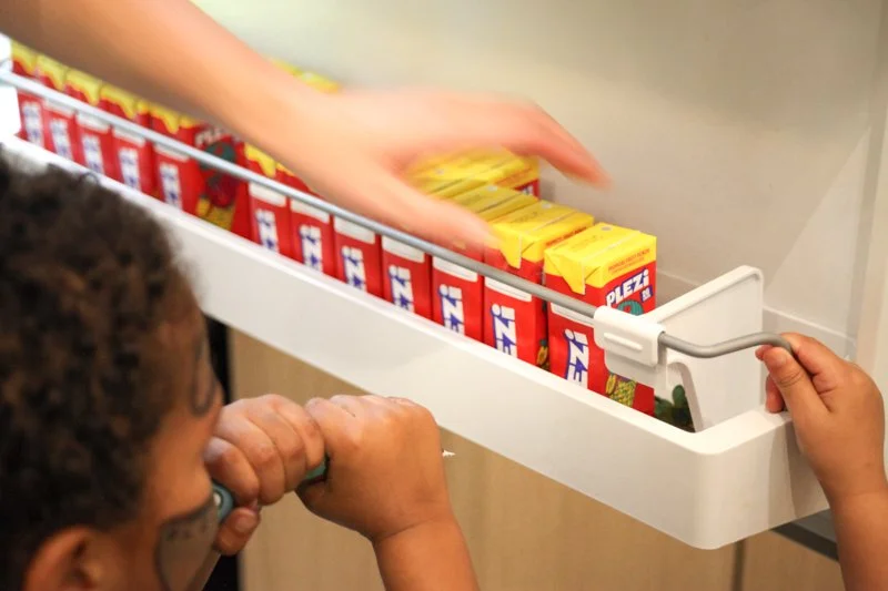 Child with curly hair looking at an open cooler with multiple small red and yellow juice boxes inside, while an adult's hand reaches for a juice box.