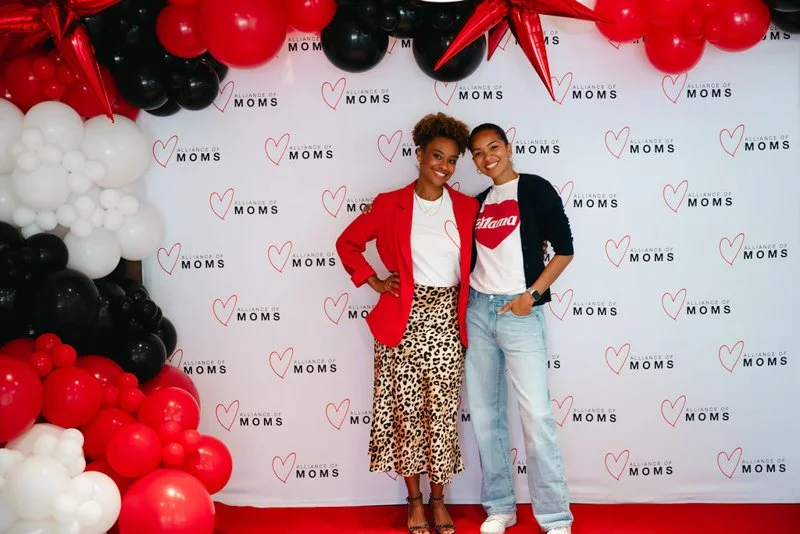 Two women standing in front of a backdrop with red and black balloon decorations, and logos that say 'Alliance of MOMS'. One woman is wearing a red blazer, white shirt, and leopard print skirt; the other is wearing a black cardigan, white t-shirt wit