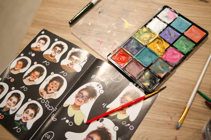 Open makeup or face paint kit with colorful paints on a wooden table, and a magazine or workbook with face painting designs and instructions. Brushes and other art tools are also visible.