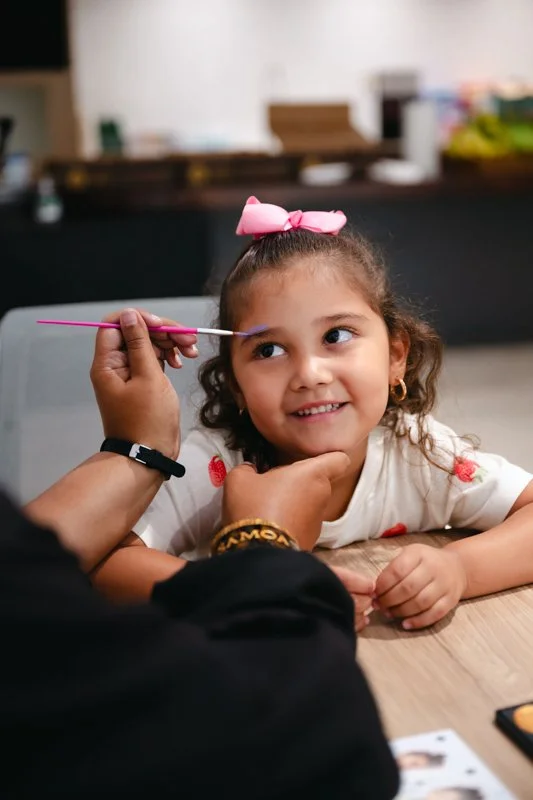 A young girl with curly hair and a pink bow on her head has her face painted with a blue line, as someone applies the paint with a brush.