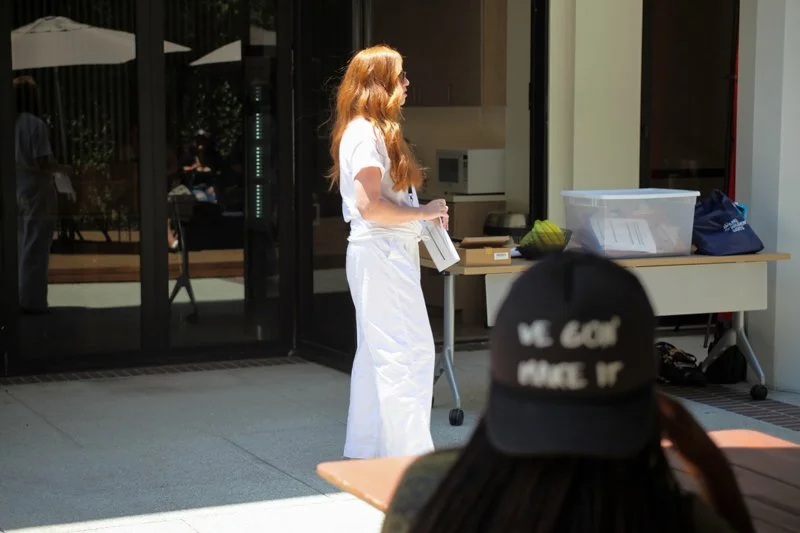 A woman with long red hair wearing white scrubs standing outside near a table with supplies, in front of a glass building. A person wearing a black hat with white text and dark hair is in the foreground.