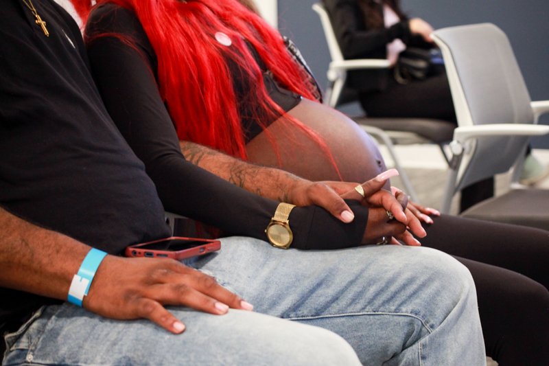 Close-up of people sitting in a waiting room, holding hands; one person has a pregnant belly.
