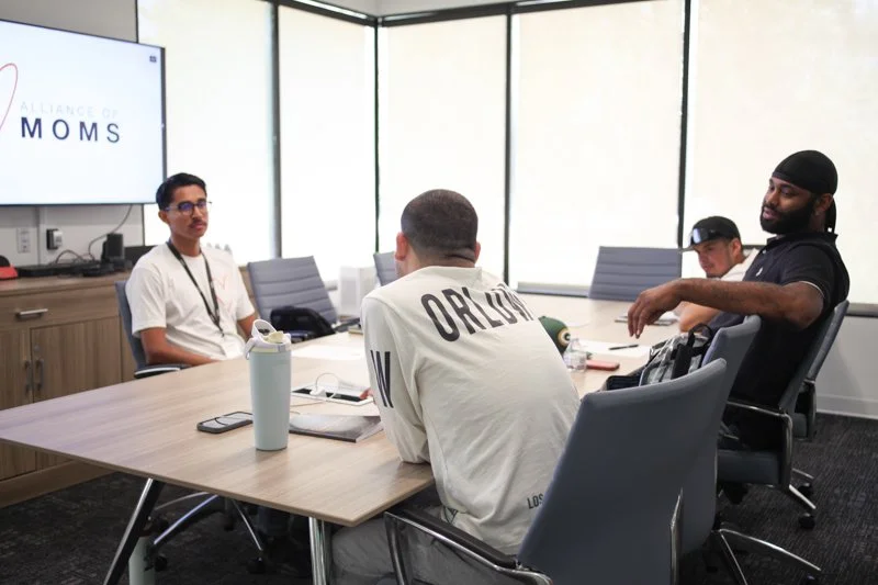 A group of four men sitting in a conference room for a meeting, with a large screen displaying the words "Alliance of Moms".